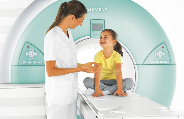Wichtig ist über die Patienten-Informationen zu erfahren, wie die Untersuchung von Kindern abläuft und was zu beachten ist. Das Bild zeigt ein Kind auf der Liege eines MRT sitzend und daneben eine Radiologische Technologin, die geduldig erklärt, was gleich bei der MRT-Untersuchung passiert.